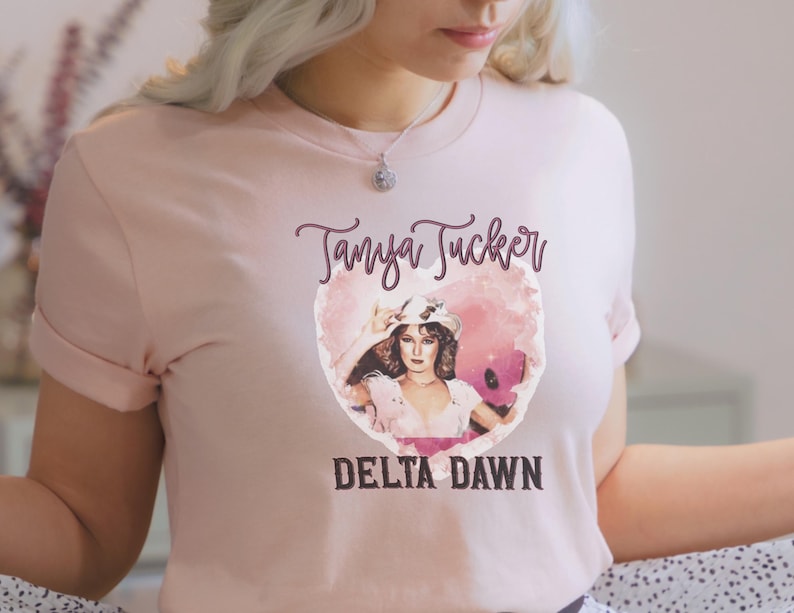 May include: Light pink t-shirt featuring a portrait of Tanya Tucker inside a heart-shaped design. The text "Tanya Tucker" is above the portrait, and "DELTA DAWN" is below. The shirt has short sleeves and a crew neck.