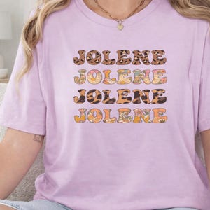 May include: A light purple short-sleeve t-shirt featuring the name JOLENE printed four times in a stacked design. The text alternates between a brown leopard print pattern and a colorful floral pattern with orange, yellow, and green flowers.