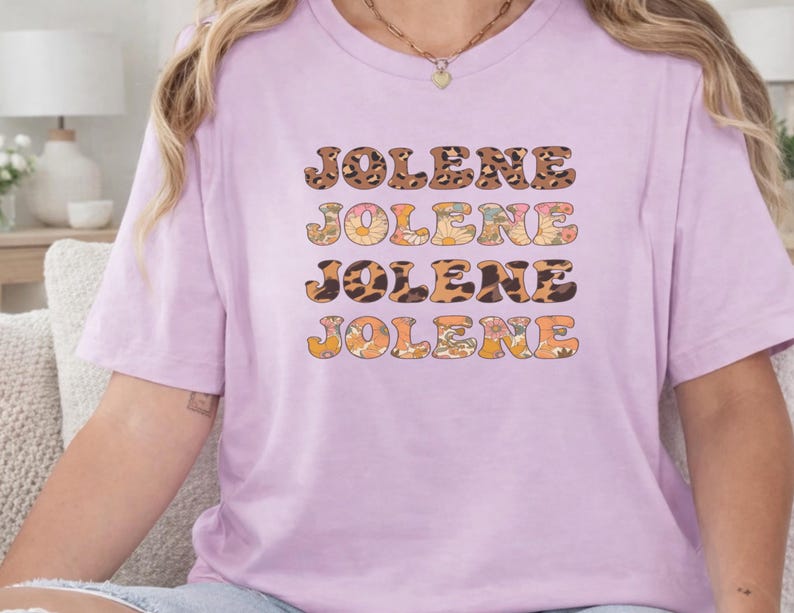 May include: A light purple short-sleeve t-shirt featuring the name JOLENE printed four times in a stacked design. The text alternates between a brown leopard print pattern and a colorful floral pattern with orange, yellow, and green flowers.