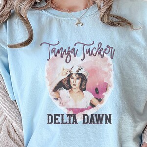 May include: Light blue t-shirt featuring a graphic of Tanya Tucker with the text "Tanya Tucker" above and "Delta Dawn" below. The graphic is in a heart shape with a pink and white watercolor effect.