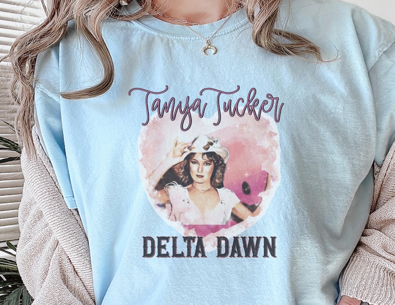 May include: Light blue t-shirt featuring a graphic of Tanya Tucker with the text "Tanya Tucker" above and "Delta Dawn" below. The graphic is in a heart shape with a pink and white watercolor effect.