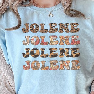 May include: A light blue t-shirt featuring the name JOLENE printed four times in a stacked design. The first and third lines have a brown and black leopard print pattern, while the second and fourth lines feature a colorful floral pattern.