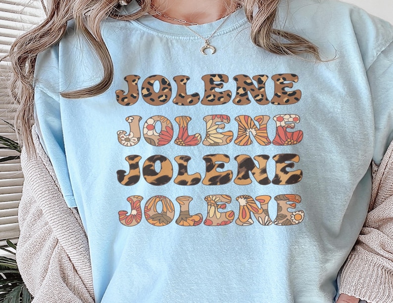 May include: A light blue t-shirt featuring the name JOLENE printed four times in a stacked design. The first and third lines have a brown and black leopard print pattern, while the second and fourth lines feature a colorful floral pattern.