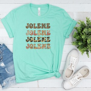 May include: A mint green short-sleeve t-shirt featuring the name JOLENE printed four times in a stacked design. Each line of text displays a different pattern, including leopard print, retro floral motifs, and brown and orange geometric shapes.