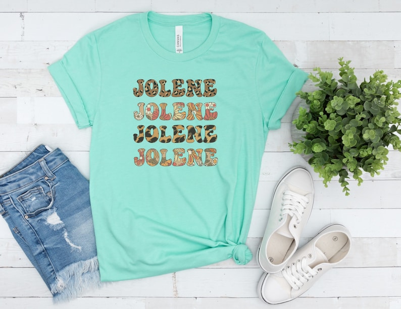 May include: A mint green short-sleeve t-shirt featuring the name JOLENE printed four times in a stacked design. Each line of text displays a different pattern, including leopard print, retro floral motifs, and brown and orange geometric shapes.