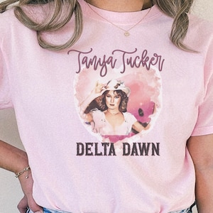 May include: Pink t-shirt featuring a graphic of Tanya Tucker with the text "Tanya Tucker" above and "Delta Dawn" below. The graphic is a vintage-style image of a woman in a hat.