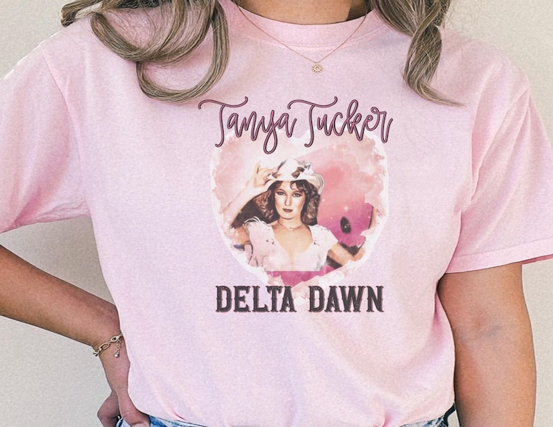 May include: Pink t-shirt featuring a graphic of Tanya Tucker with the text "Tanya Tucker" above and "Delta Dawn" below. The graphic is a vintage-style image of a woman in a hat.
