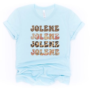 May include: A light blue short-sleeve t-shirt featuring the name JOLENE printed four times in a stacked design. The text alternates between a brown leopard print pattern and a multi-colored floral pattern with orange, red, and white flowers.
