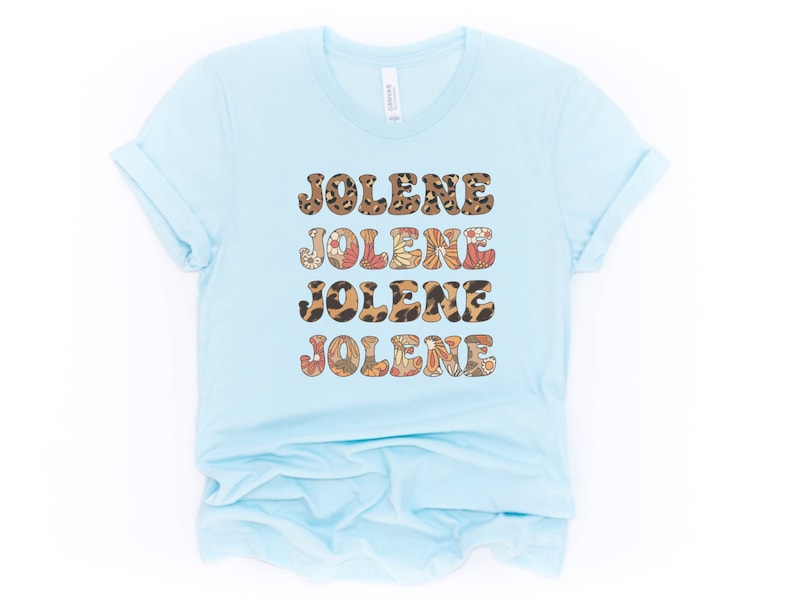 May include: A light blue short-sleeve t-shirt featuring the name JOLENE printed four times in a stacked design. The text alternates between a brown leopard print pattern and a multi-colored floral pattern with orange, red, and white flowers.