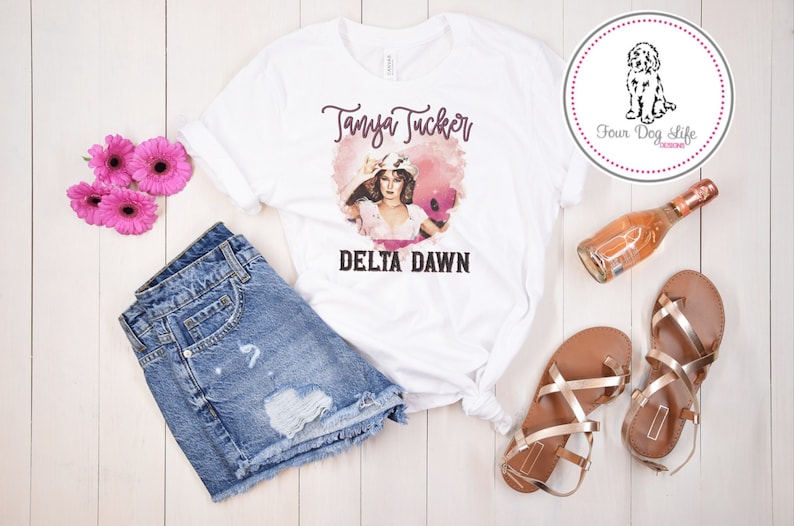 May include: White t-shirt featuring a graphic of Tanya Tucker and the words "Delta Dawn." The shirt is styled with denim shorts, gold sandals, and pink flowers. A bottle of ros&eacute; and a dog logo are also present.
