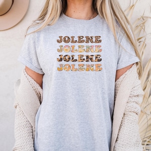 May include: A light gray short-sleeved t-shirt featuring the name JOLENE printed four times in a stacked, wavy font. Each line of text displays a different pattern, including leopard print, floral motifs, and solid orange, creating a layered effect.