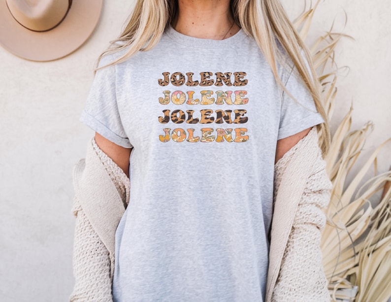 May include: A light gray short-sleeved t-shirt featuring the name JOLENE printed four times in a stacked, wavy font. Each line of text displays a different pattern, including leopard print, floral motifs, and solid orange, creating a layered effect.