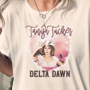 May include: White t-shirt featuring a graphic of Tanya Tucker with the text "Tanya Tucker" in a cursive font above the text "Delta Dawn" in a bold font. The graphic is set within a pink heart shape.