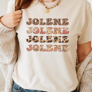 May include: A cream-coloured t-shirt with the name "JOLENE" printed four times in a stacked arrangement. The name is rendered in various patterns, including leopard print and floral designs in brown, orange, and cream hues.