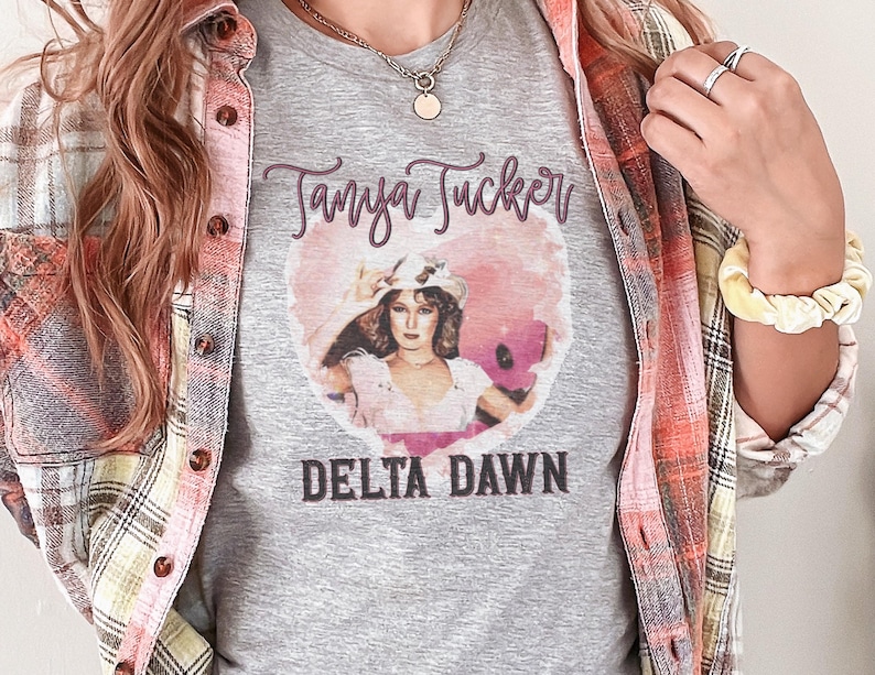 May include: A gray t-shirt featuring a portrait of Tanya Tucker inside a pink heart, with the text "Tanya Tucker" above and "Delta Dawn" below. The person is wearing a plaid shirt.