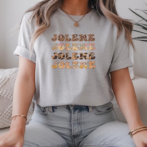 May include: A light gray short-sleeved t-shirt featuring the name JOLENE printed four times in a stacked design. Each line of text displays a different pattern, including leopard print, floral motifs, and solid orange, with a crew neckline.