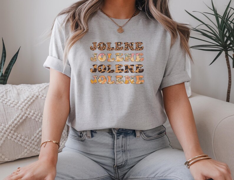 May include: A light gray short-sleeved t-shirt featuring the name JOLENE printed four times in a stacked design. Each line of text displays a different pattern, including leopard print, floral motifs, and solid orange, with a crew neckline.