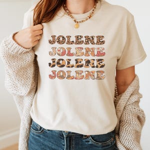 May include: A cream-colored short-sleeve t-shirt featuring the name JOLENE printed four times in a stacked design. The first and third lines have a brown leopard print pattern, while the second and fourth lines feature a colorful floral pattern.