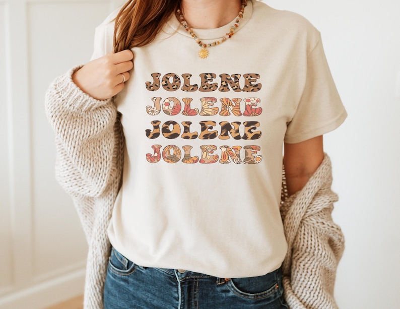 May include: A cream-colored short-sleeve t-shirt featuring the name JOLENE printed four times in a stacked design. The first and third lines have a brown leopard print pattern, while the second and fourth lines feature a colorful floral pattern.