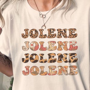 May include: A white short-sleeved t-shirt featuring the name JOLENE printed four times in a stacked design. The first and third lines have a brown and black leopard print pattern, while the second and fourth lines feature a floral pattern.