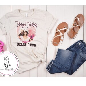 May include: A cream-colored t-shirt featuring a graphic of Tanya Tucker and the words "DELTA DAWN." The image is accompanied by blue jeans, gold sandals, and pink flowers. A circular logo with a dog illustration is also present.
