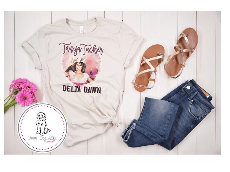 May include: A cream-colored t-shirt featuring a graphic of Tanya Tucker and the words "DELTA DAWN." The image is accompanied by blue jeans, gold sandals, and pink flowers. A circular logo with a dog illustration is also present.