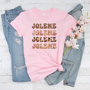 May include: A light pink short-sleeved t-shirt featuring the name JOLENE printed four times in a stacked design. The text uses alternating patterns of brown leopard print and a multi-colored floral motif with orange, red, and white accents.