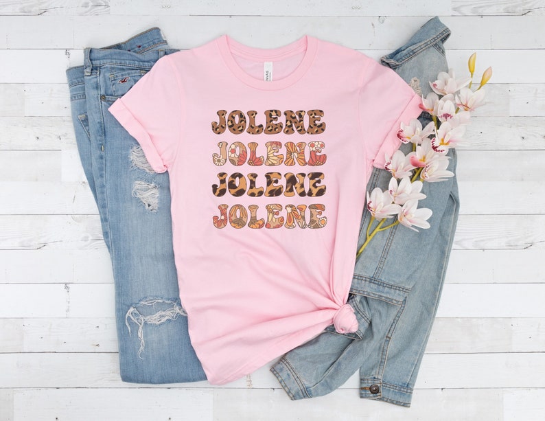 May include: A light pink short-sleeved t-shirt featuring the name JOLENE printed four times in a stacked design. The text uses alternating patterns of brown leopard print and a multi-colored floral motif with orange, red, and white accents.