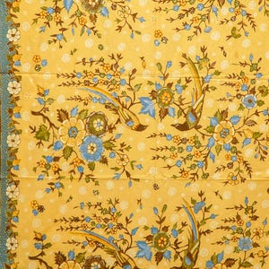 Puede incluir: Tela amarilla con un patrón floral y de pájaros repetido. El diseño presenta flores azules, marrones y amarillas con pájaros azules sobre un fondo amarillo. Un borde azul y blanco enmarca la tela.