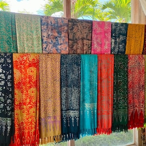 May include: A collection of colorful patterned scarves with fringe hanging from the bottom. The scarves are displayed on a wooden rod against a window.