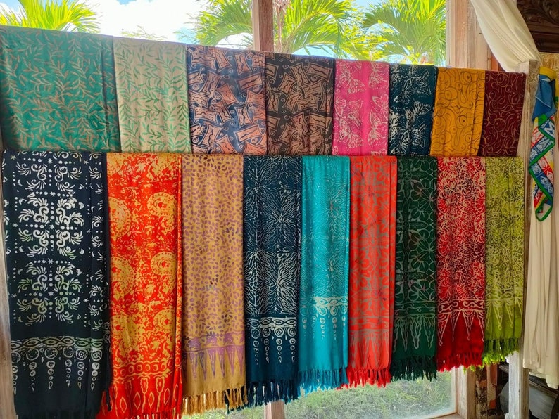 May include: A collection of colorful patterned scarves with fringe hanging from the bottom. The scarves are displayed on a wooden rod against a window.