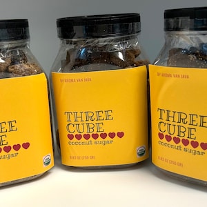 May include: Three clear plastic jars filled with dark brown coconut sugar cubes. Each jar features a yellow label with the text "THREE CUBE coconut sugar" and a row of red hearts. The labels also state "BY AROMA VAN JAVA" and "8.83 OZ (250 GR)".