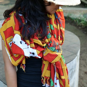 May include: A woman wearing a black top and a red, yellow, and white patterned scarf with playing cards and the text "To Las Vegas". She is wearing a white straw hat with a black ribbon.
