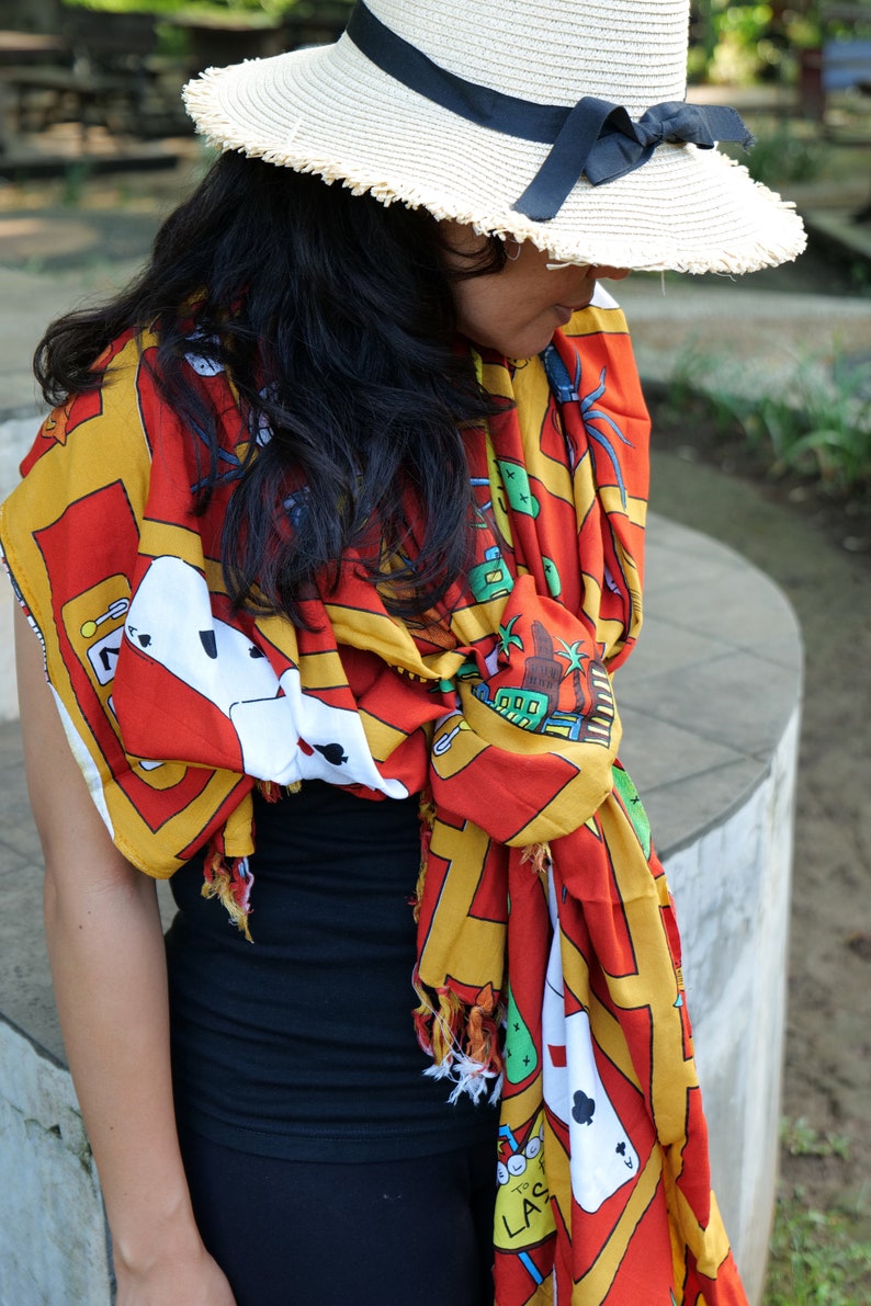 May include: A woman wearing a black top and a red, yellow, and white patterned scarf with playing cards and the text "To Las Vegas". She is wearing a white straw hat with a black ribbon.