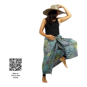 May include: A woman wearing a black tank top and a patterned sarong. She is wearing a wide-brimmed straw hat and has her hair pulled back. The sarong is tied in a knot at the waist and has a flowing design.