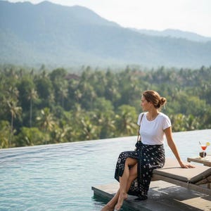 Puede incluir: Una persona con una camiseta blanca y un pareo con estampado en blanco y negro se sienta junto a una piscina infinita. El fondo presenta exuberantes árboles verdes y montañas, con un cóctel en una mesa cercana.