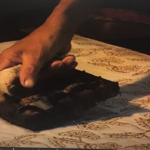 May include: A person using a wooden block with a carved design to stamp a pattern onto a white fabric. The fabric has a floral pattern already printed on it.