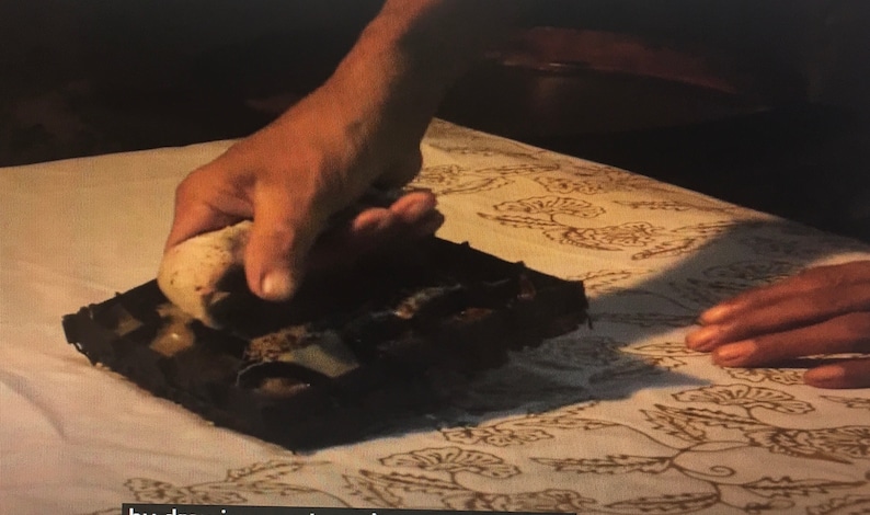 May include: A person using a wooden block with a carved design to stamp a pattern onto a white fabric. The fabric has a floral pattern already printed on it.