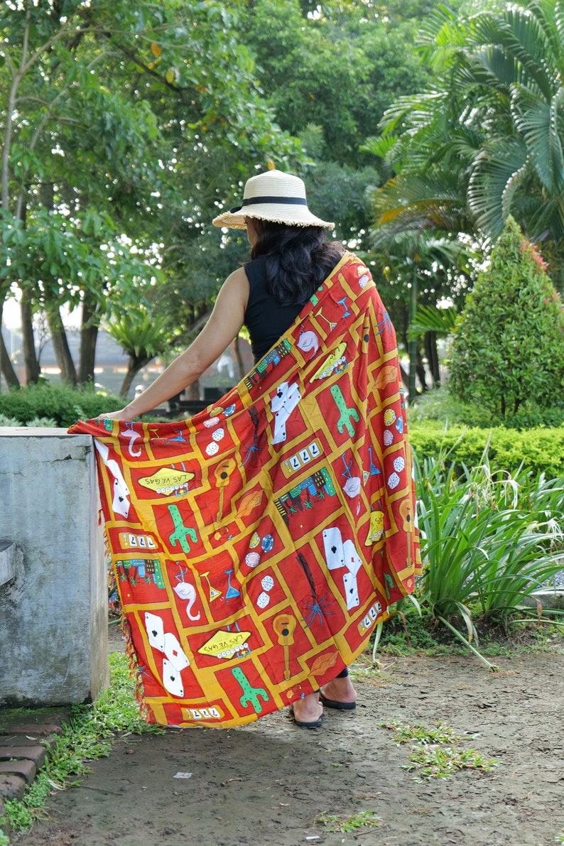 May include: A woman wearing a straw hat and a red, yellow, and blue patterned shawl with a Las Vegas theme. The shawl features images of playing cards, dice, guitars, and cacti.