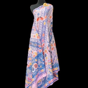 Op de afbeelding: Een sjaal met bloemmotieven gedrapeerd over een mannequin. De sjaal heeft een lichtroze achtergrond met blauwe, gele en oranje bloempatronen. De randen zijn afgewerkt met een blauwe rand. Een goudkleurige broche siert de schouder van de mannequin.