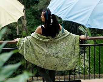 May include: A woman wearing a green and white patterned shawl stands on a balcony overlooking a lush green forest. She is wearing a black tank top and dark green pants. A blue and white umbrella is behind her.