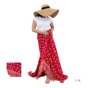 Op de afbeelding: Een rode sarong met een gouden bladerenpatroon en franjes. De sarong is om de taille gewikkeld. Een grote strohoed met een zwarte band wordt op het hoofd gedragen. De persoon draagt een wit T-shirt.