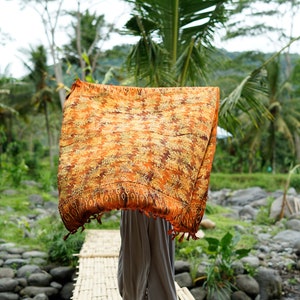 May include: A person walks across a bamboo bridge wearing a long, flowing orange and brown patterned scarf. The bridge is over a small stream with rocks on the banks. Lush green foliage surrounds the bridge.