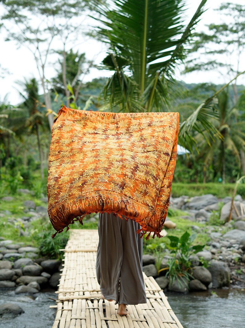 May include: A person walks across a bamboo bridge wearing a long, flowing orange and brown patterned scarf. The bridge is over a small stream with rocks on the banks. Lush green foliage surrounds the bridge.
