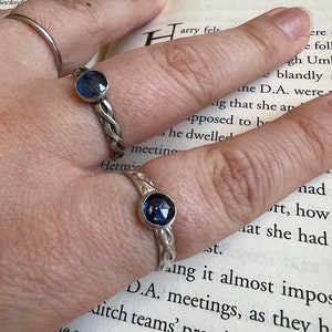 Peut inclure: Deux bagues en argent avec un motif de bande tressée et une pierre précieuse bleu foncé. Une bague est sur un doigt, l'autre est légèrement floue en arrière-plan. Les bagues sont posées sur un livre ouvert.