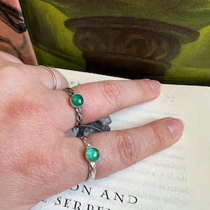Peut inclure: Deux bagues en argent avec une pierre précieuse verte ronde et vibrante sertie dans une monture décorative. Les bagues sont présentées sur une main, avec un livre en arrière-plan. Les bagues sont un bijou.