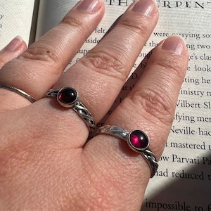 May include: Two silver rings with a braided design and a round, deep red gemstone. The rings are on a hand, with a book in the background. The text on the book reads "THE LION AND THE SERPENT".