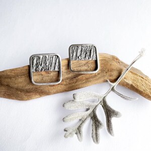 May include: A pair of silver square earrings with a textured design. The earrings are on a piece of driftwood with a gray leaf.