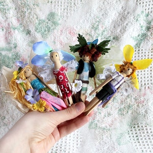 May include: Four handmade wooden dolls dressed as fairies with colourful wings, flowers, and fabric clothing. The dolls are held in a hand against a white crocheted background.