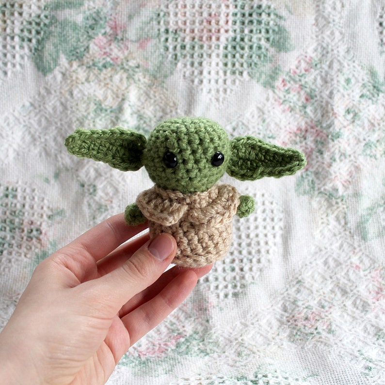 Pu&ograve; includere: Un giocattolo di peluche verde e marrone all'uncinetto che ricorda un personaggio del franchise di Star Wars. Il giocattolo ha occhi grandi e orecchie piccole.